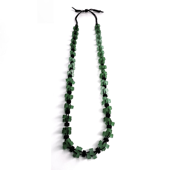 A long, hypoallergenic Verona Long Necklace handcrafted in Paris by Alex + Svet, featuring interconnected small green cross shapes that transition from darker to lighter hues towards the center. The necklace is tied with black cords at the ends, making it an elegant addition to any ensemble.