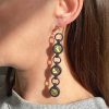 Close-up of a person with straight brown hair wearing Elsiem's Vibe 3 Drop Earrings—statement pieces featuring four connected black rings with green stones, 3D printed and finished with gold filled ear posts. They're paired with a light-colored top.