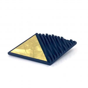 A black, triangular prism with one gold-colored face in brushed finished lacquered brass and evenly spaced black ridges on the other faces is displayed on a white background. The unique geometric design creates an impression of modern elegance, reminiscent of the Elsiem Vibe Wave Fascia Brooch.