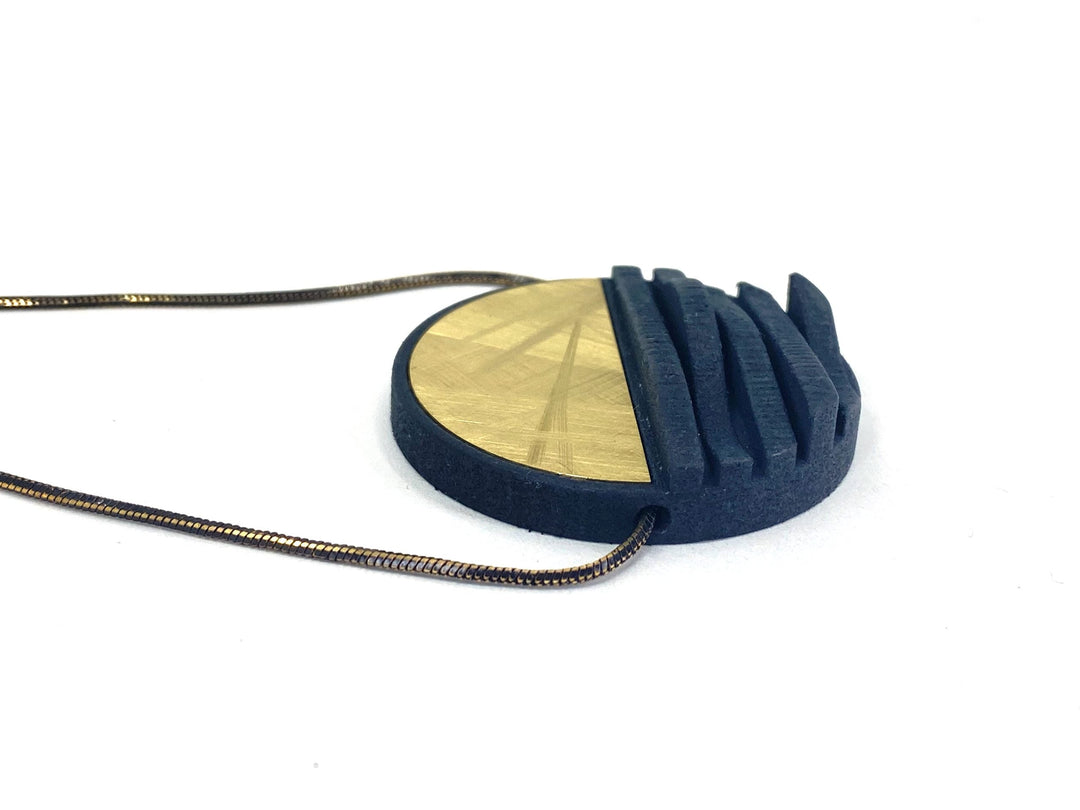 The Elsiem Vibe Wave Circle Fascia Necklace features a modern round pendant split into half-gold, half-dark blue. The blue side has raised curved ridges, while the gold side shows an etched geometric pattern, all on a slim metallic chain.