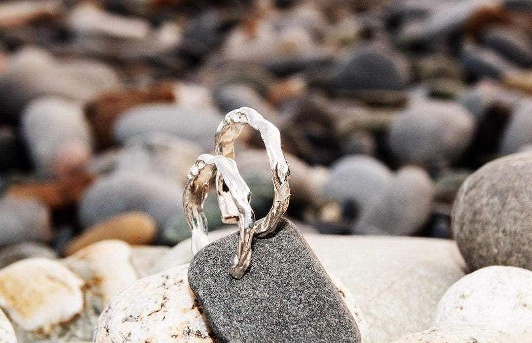 Two textured silver hoop earrings from Ava Jane Jewellery’s "Waves of The Irish Sea" collection rest on a smooth, dark stone among pebbles, evoking natural beauty with a softly blurred background of rocks and stones.