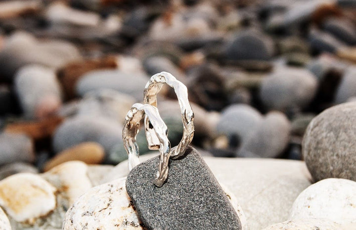 Two textured silver hoop earrings from Ava Jane Jewellery’s "Waves of The Irish Sea" collection rest on a smooth, dark stone among pebbles, evoking natural beauty with a softly blurred background of rocks and stones.
