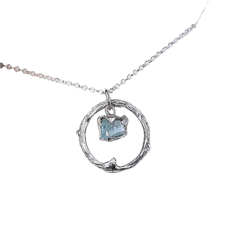 A close-up image of the Raw Aquamarine Halo Pendant by Caroline Stokesberry-lee. The sterling silver necklace with a circular pendant showcases a small, raw aquamarine gemstone inside the circle. The one-of-a-kind necklace is displayed in an open beige jewelry box.