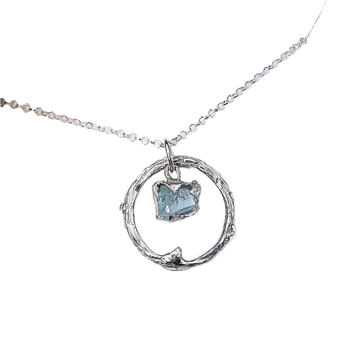 A close-up image of the Raw Aquamarine Halo Pendant by Caroline Stokesberry-lee. The sterling silver necklace with a circular pendant showcases a small, raw aquamarine gemstone inside the circle. The one-of-a-kind necklace is displayed in an open beige jewelry box.