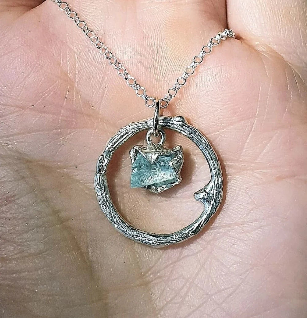 Close-up of a person's hand holding a sterling silver necklace with a circular pendant. The Raw Aquamarine Halo Pendant by Caroline Stokesberry-lee features a small, raw aquamarine crystal at its center. The fingers are gently cradling the one-of-a-kind necklace, highlighting its intricate design and texture.
