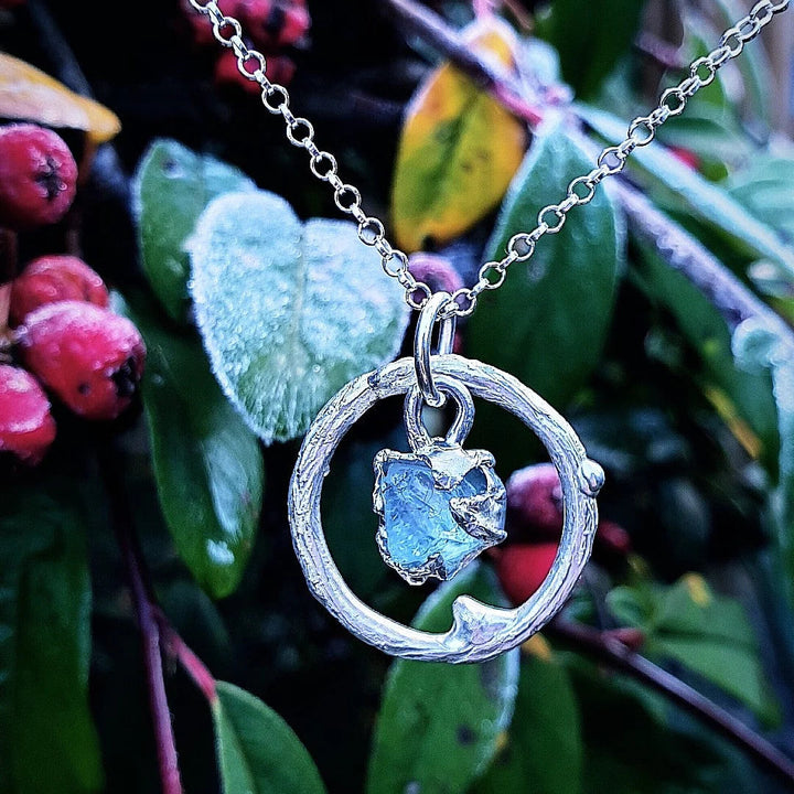 A sterling silver Caroline Stokesberry-lee Raw Aquamarine Halo Pendant featuring a circular design with a small, rough Raw Aquamarine stone in the center hangs against a backdrop of frosty green leaves and red berries. The intricate details of this one-of-a-kind pendant and the natural background create a striking visual contrast.