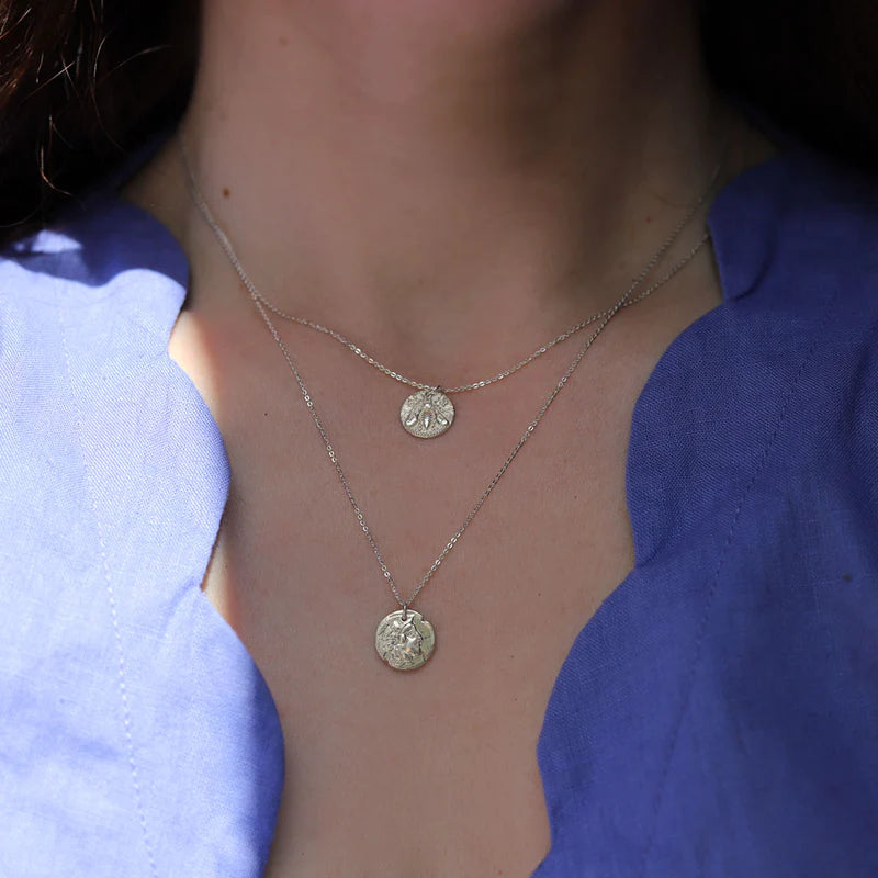 Close-up of a person wearing two sterling silver necklaces with round, ornate pendants. One features an Athena Coin Necklace Silver by Andrea Mears Jewellery, the other a symbolic coin necklace. The person is dressed in a blue top with a wavy neckline. The pendants have intricate designs that catch the light, with the background blurred to focus on the jewelry and neckline.