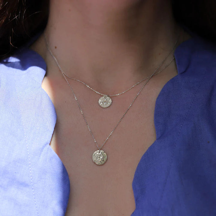 Close-up of a person wearing two sterling silver necklaces with round, ornate pendants. One features an Athena Coin Necklace Silver by Andrea Mears Jewellery, the other a symbolic coin necklace. The person is dressed in a blue top with a wavy neckline. The pendants have intricate designs that catch the light, with the background blurred to focus on the jewelry and neckline.