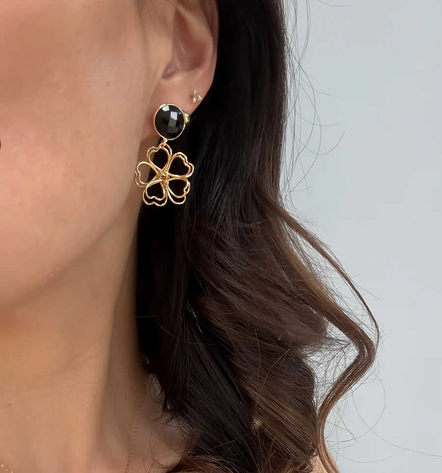 A close-up of a woman's ear wearing Aria V's Black Daisy Earrings—gold with a black stud and four linked heart-shaped charms. She has wavy brown hair set against a plain background.