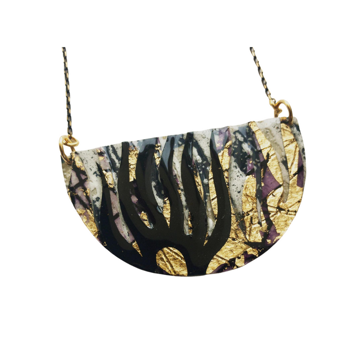 Presenting the Bláth Batik Textile Necklace by Rothlú, this exquisite piece features a half-circle pendant with unique textures and an abstract design in black, gold, and purple accents. The pendant is finished with eco resin and is elegantly attached to a chain by two gold rings on either side.