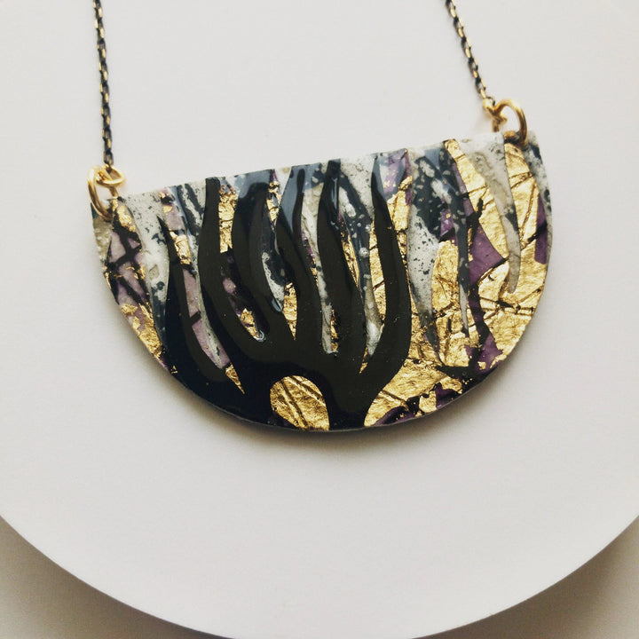 A close-up of the Bláth Batik Textile Necklace in Dove/Black/Gold by Rothlú. The semi-circular pendant on a chain features a unique textured design with black, gold, and dove colors. The black forms resemble tree branches, while the gold and dove patterns add a textured, artistic look. This Rothlú batik necklace is finished with eco resin.