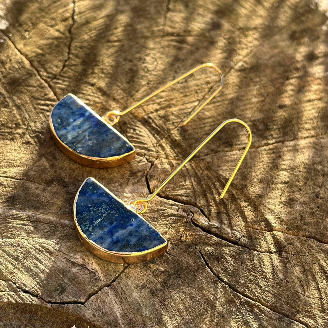 The Blue Moon Lapis Earrings by Kybalion Jewellery, featuring gold-plated hooks and semi-circle Lapis Lazuli pendants, elegantly rest on a textured wooden surface. Sunlight casts radiant patterns, enhancing the handmade earrings' reflective surfaces.