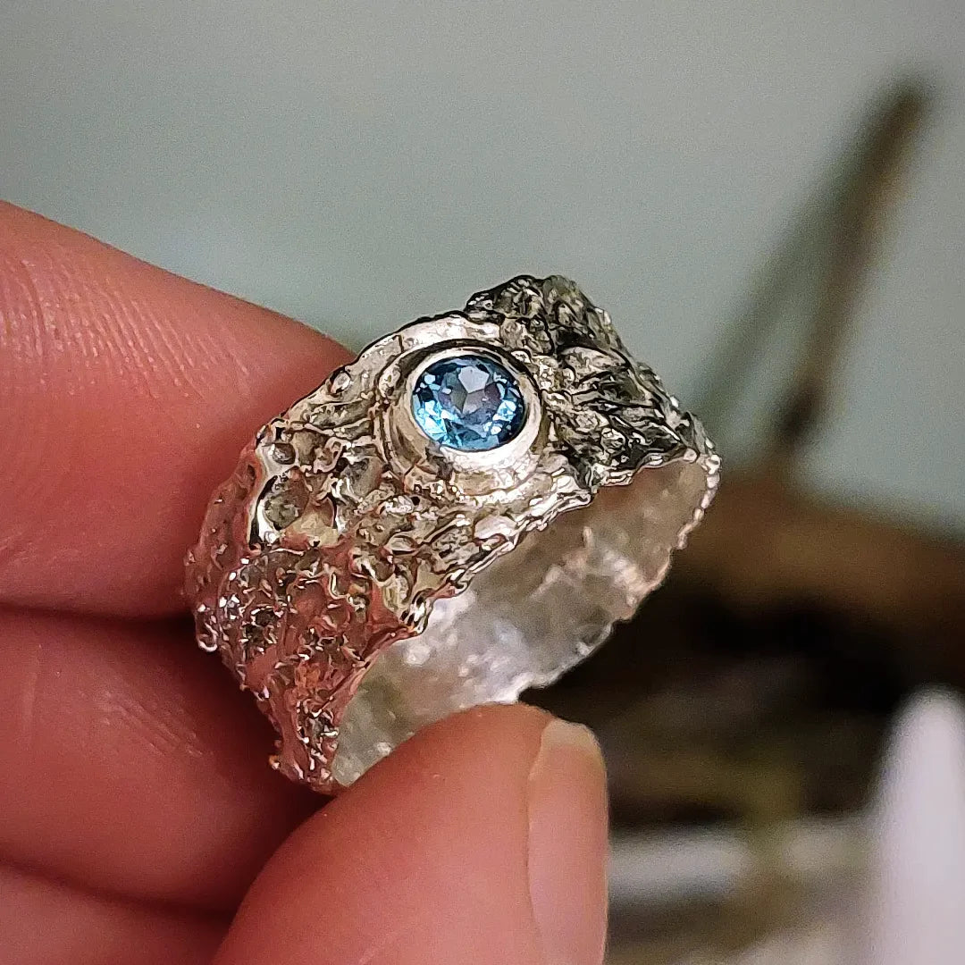A hand holds the Caroline Stokesberry-lee "That Place Beyond Silver Slim Band Ring with Gemstone," featuring a textured sterling silver band and a round blue gemstone at its center.
