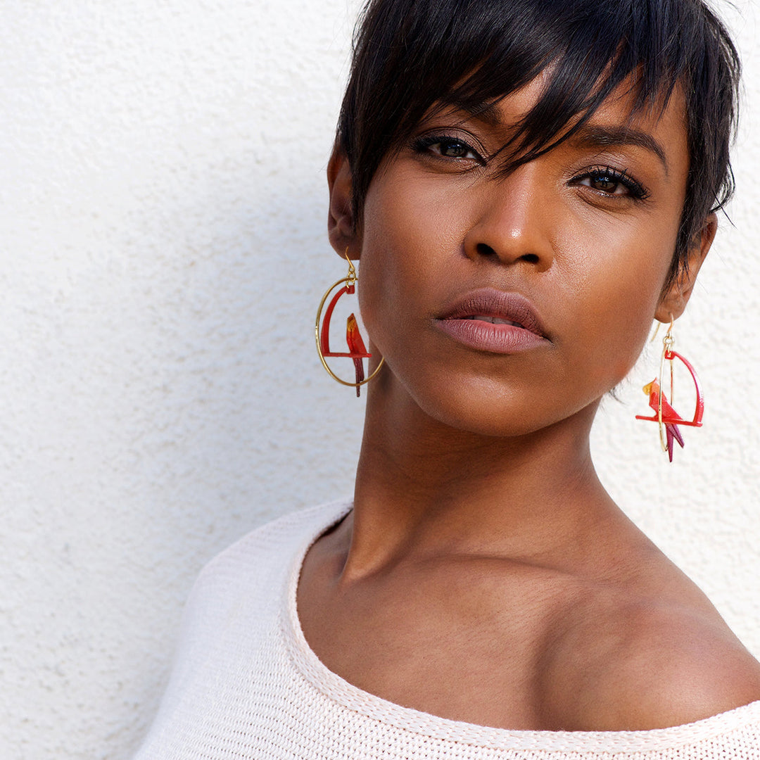 A woman with short, dark hair showcases Tochu-De’s Fuchsia Origami Parrot Earrings, wearing a light off-the-shoulder top as she poses confidently against a white textured wall.