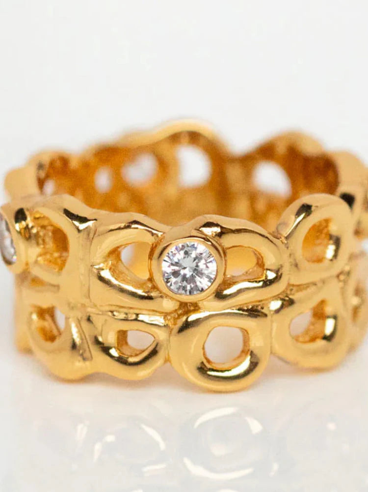 The Shyla Bordeaux Ring is a handmade gold ring with an intricate openwork design, featuring loops and round, clear gemstones evenly spaced around the hypoallergenic band—an elegant jewellery piece.