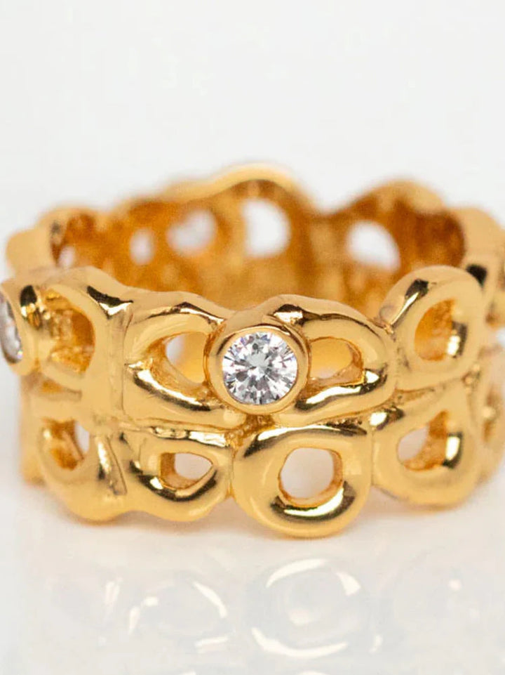 The Shyla Bordeaux Ring is a handmade gold ring with an intricate openwork design, featuring loops and round, clear gemstones evenly spaced around the hypoallergenic band—an elegant jewellery piece.