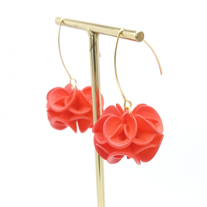 A delicate pair of Tochu-De Orange Rose Earrings featuring vibrant red flower-shaped pendants hanging from thin, gold hooks. The 3D printed jewelry is displayed on a sleek, gold stand against a plain white background.