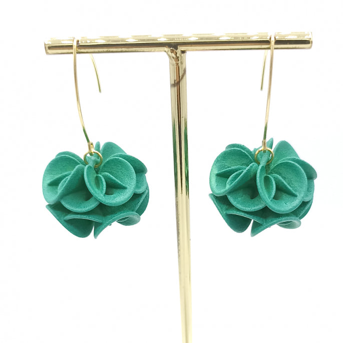 A pair of Tochu-De Emerald Rose Earrings with turquoise blue fabric flowers hangs from a gold display stand. The flowers have a ruffled texture, creating a voluminous, charming appearance. The hoop portion of each earring loops through the center of each flower, combining elegance with a whimsical touch.