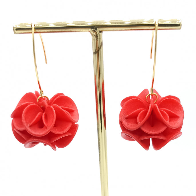A pair of colorful fantasy earrings featuring intricate red fabric flowers hanging from thin gold hooks. The Red Rose Earrings by Tochu-De are suspended from a gold display stand against a white background, creating an enchanting and elegant aesthetic.