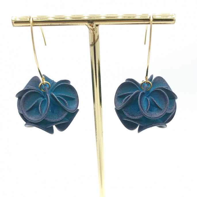 A pair of Petrol Rose Earrings by Tochu-De featuring intricate, dark blue flower designs made from leather hangs from a simple, elegant golden stand. The flowers have layered petals, giving them a full, textured appearance. The fantasy earrings are suspended from gold hooks.