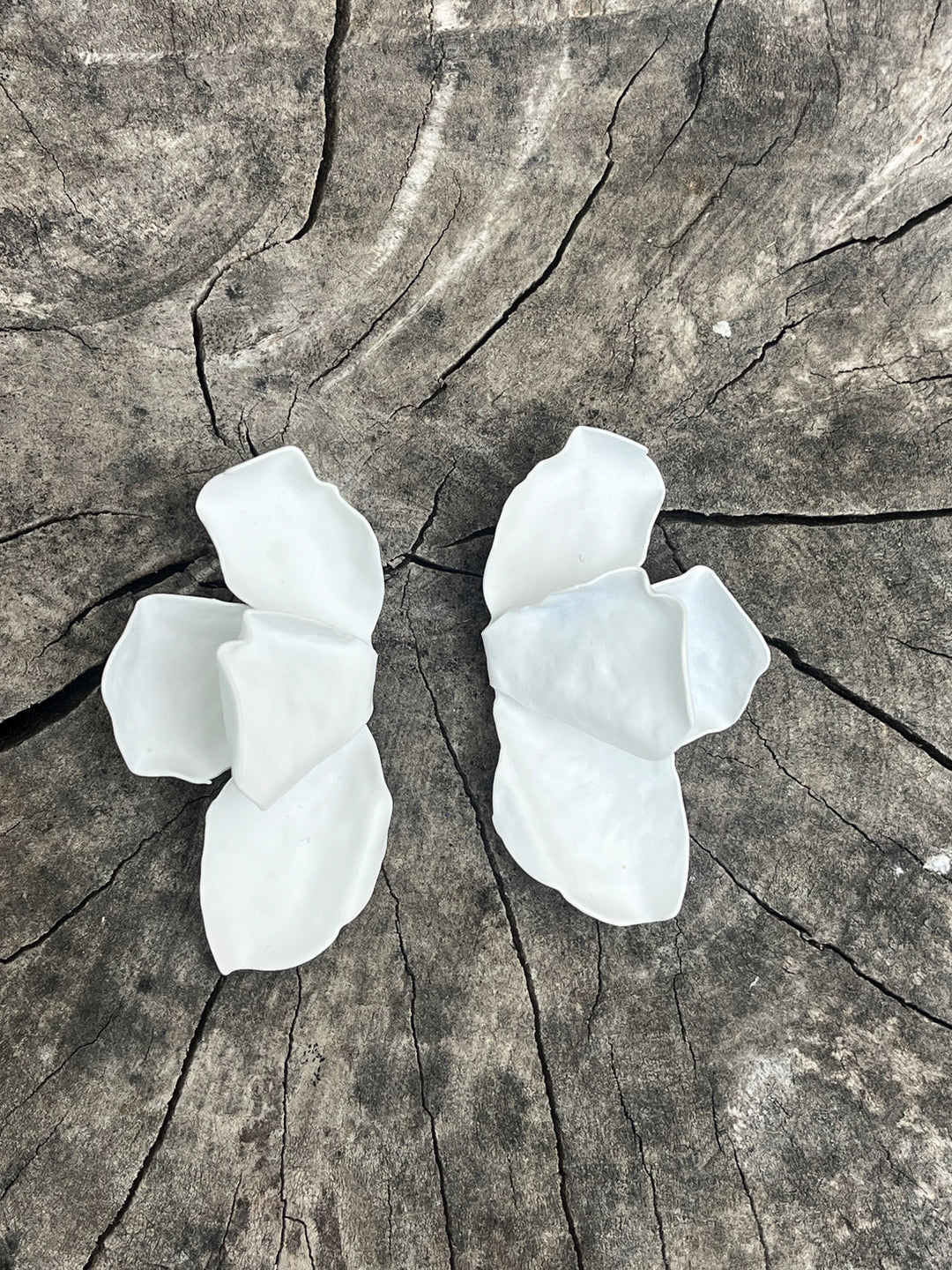 The Bougainvillea Earrings by Kybalion Jewellery, shaped like flower petals and made from white ceramic, are symmetrically displayed on a textured, cracked wooden surface. Handmade with care and nickel-free, they offer a delicate and natural look.