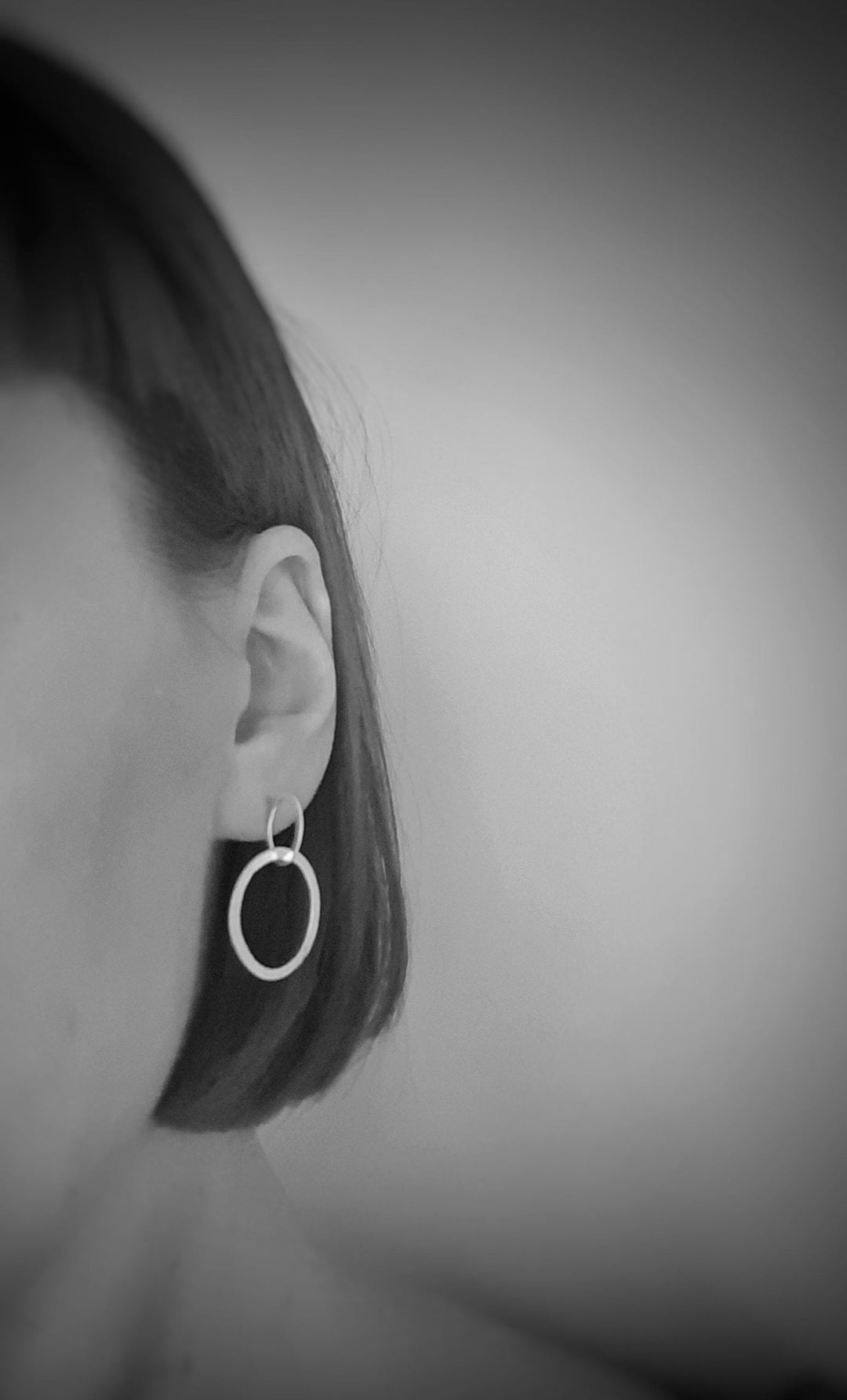 Black and white image showing the side profile of a person with short hair, emphasizing a circular, hand-forged sterling silver earring. The focus is on the ear and the earring, Carran Drops by Lynsey de Burca, with a blurred background. The overall tone is minimalist and elegant.