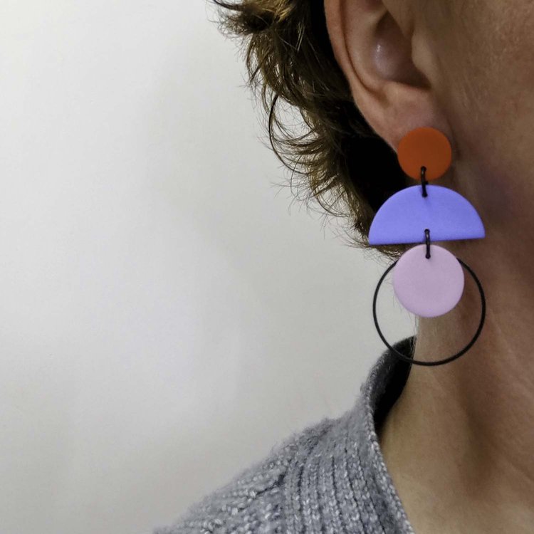Close-up of a person's ear wearing Culu Design's Cascade Red Blue & Lilac Earrings—handmade polymer clay with red, blue, and lilac geometric shapes on a black hoop. The person has short brown hair and a textured gray top against a light background.