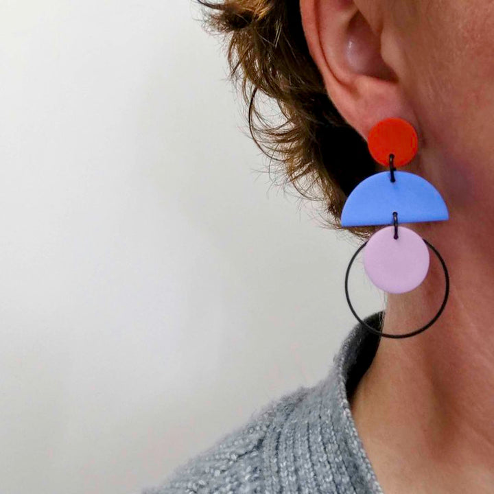 Close-up of a person's ear wearing Culu Design's Cascade Red Blue & Lilac Earrings—handmade polymer clay with red, blue, and lilac geometric shapes on a black hoop. The person has short brown hair and a textured gray top against a light background.