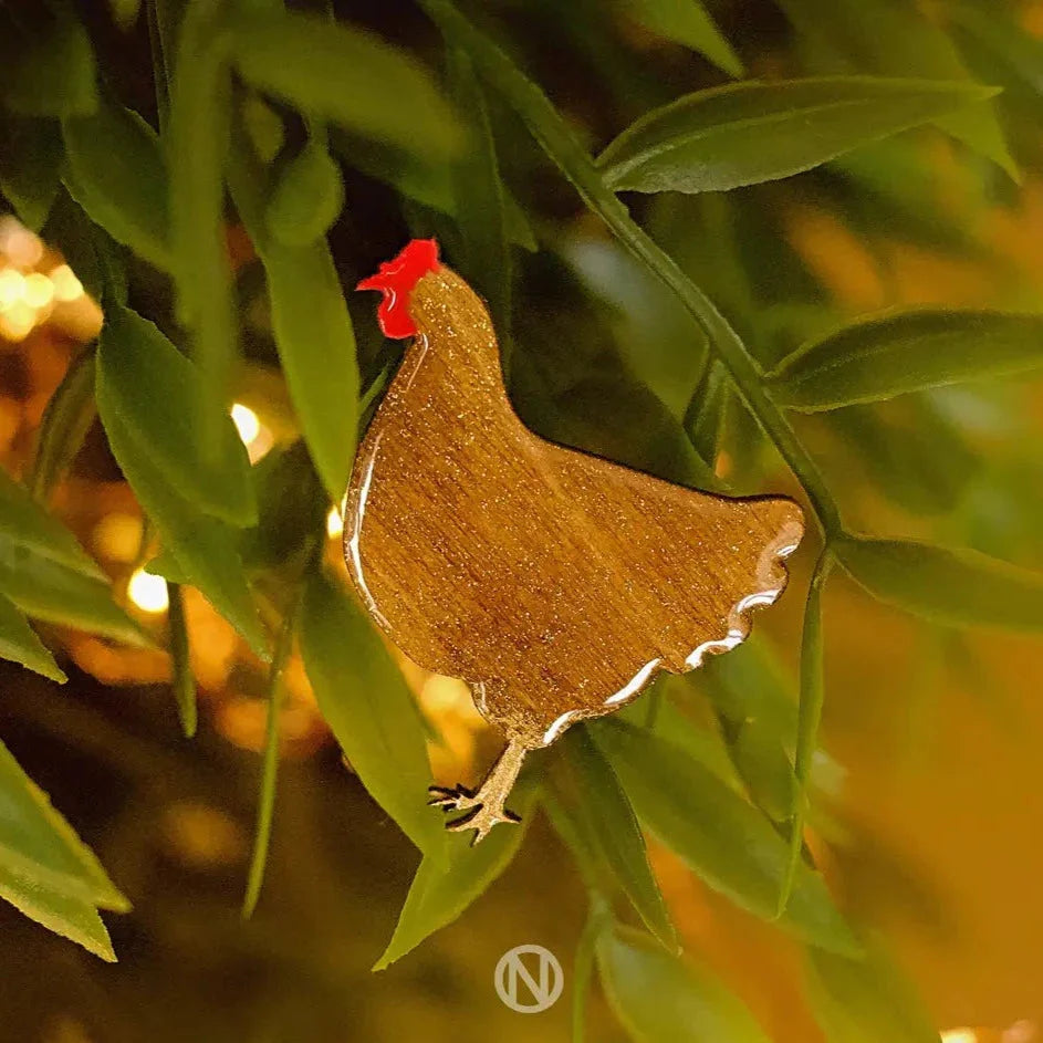 The Chicken Pin Brooch Gold by Naoi features a metallic chicken with a red comb atop green leaves, secured with a brass butterfly clasp, all set against a warm golden background for a cozy touch.
