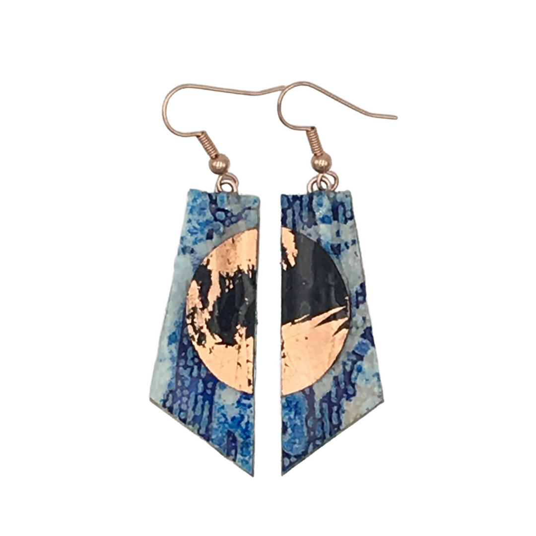 A pair of Rothlú Cora Batik Textile Earrings in Blue/Black/Rose Gold featuring a copper circular design split into two parts. The unique texture of the blue and white marbled background accents these triangular earrings, which have slightly curved bottom edges and hang from hypo-allergenic rose gold plated hooks.