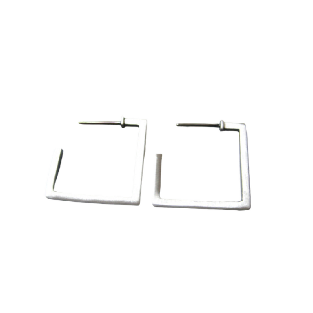 A pair of oxidized silver, square-shaped hoop earrings with open ends lie flat on a white surface. The minimalist design features thin, architectural lines, and the earrings are positioned side by side, slightly apart. These are the Cub Square Earrings by MarCucurella.