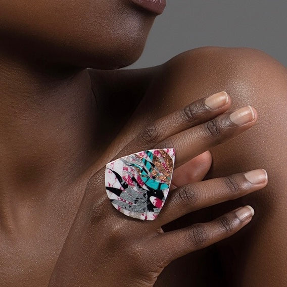 A person wears a striking, large teardrop-shaped adjustable ring on their finger. The Díog Ring Pink/Black/Silver/Blue/Rose-Gold by Rothlú features a colorful abstract pattern with shades of pink, turquoise, black, and gold on a white background. Made from eco resin jewelry with hypoallergenic silver-plated material, it beautifully complements their dark skin tone against the plain gray background.