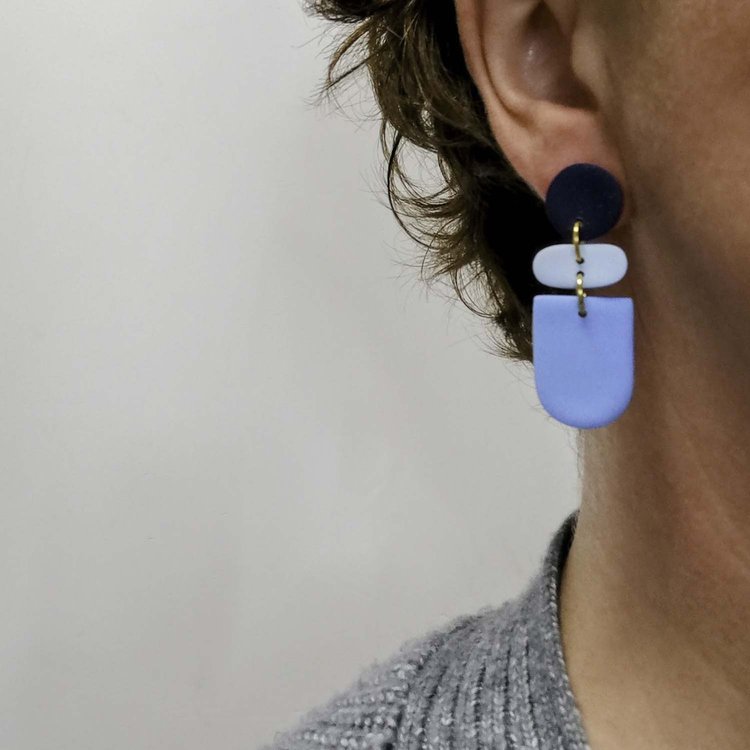 Close-up of a person with short, wavy brown hair wearing Culu Design’s Drift Midnight Blue Earrings—handmade jewelry with navy, light blue, and blue geometric shapes—paired with a grey textured top against a light background.