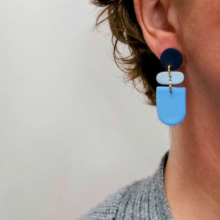 Close-up of a person with short, wavy brown hair wearing Culu Design’s Drift Midnight Blue Earrings—handmade jewelry with navy, light blue, and blue geometric shapes—paired with a grey textured top against a light background.