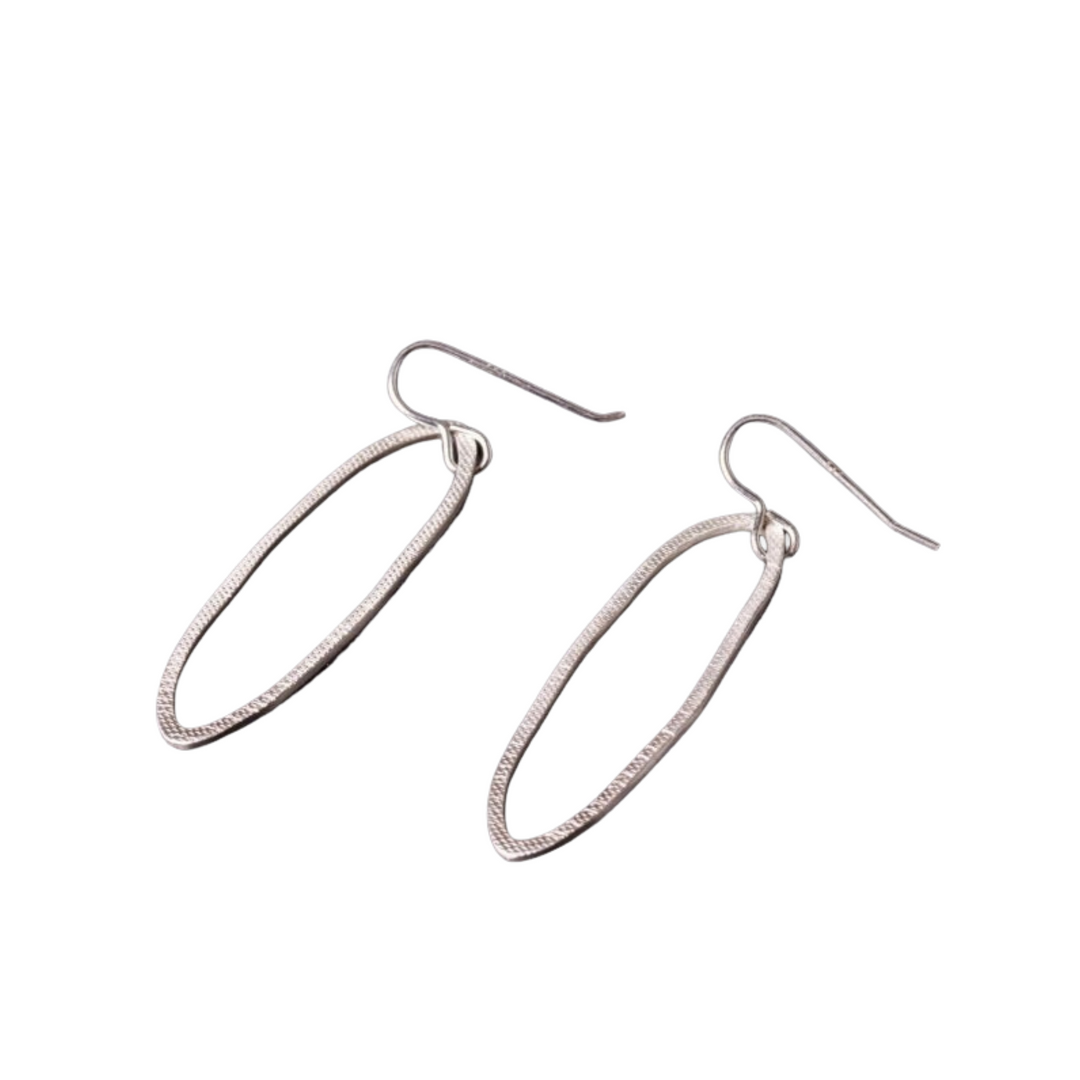 A pair of Lynsey de Burca Drift Drop Earrings with hook attachments, resting against a white background. The hand-forged earrings feature a sleek, elongated oval design with a slight texture along the edges, reminiscent of fishing net rope.