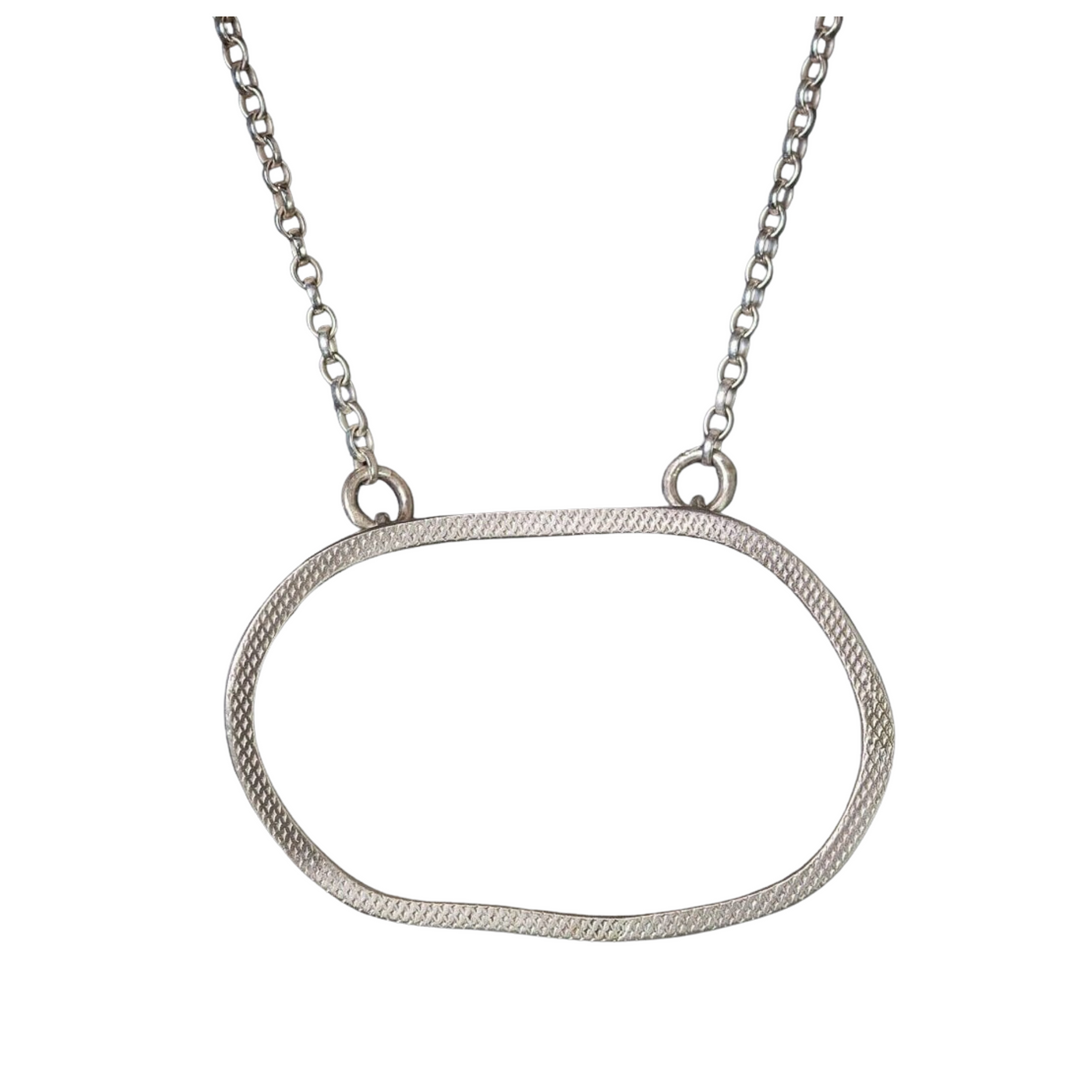 A Lynsey de Burca Drift Pendant Silver, a hand-forged silver necklace with a thin, irregularly-shaped oval pendant suspended from a chain with small round links. The textured sterling silver pendant adds subtle detail to its minimalist design. Its simple elegance makes it an understated accessory.