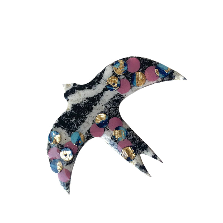 The Éan Bird Batik Textile Brooch by Rothlú features a stylized bird with black and white stripes, accented with pink, blue, and gold confetti spots on white, handmade with an eco resin finish.