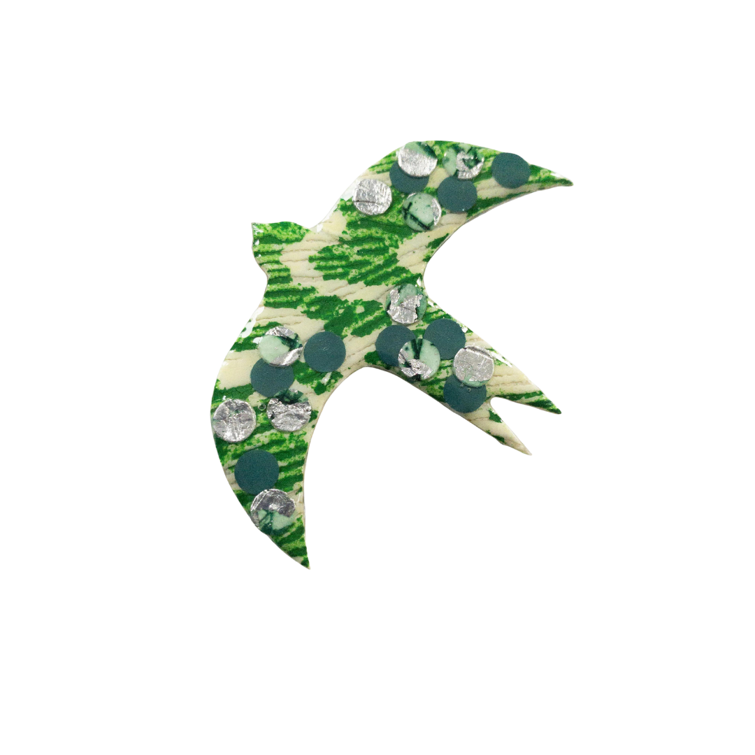 A decorative, green and white bird-shaped pendant with circular, metallic elements embedded on its surface, set against a plain, black background. The bird is in an abstract design with open wings and a pointed beak, showcasing a unique texture through an eco resin finish. This elegant piece is the Éan Bird Batik Textile Brooch by Rothlú.