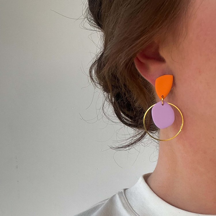 Close-up of a person wearing Culu Design's Eden Orange & Lilac Earrings—colorful polymer clay dangles with an orange top, lavender circle, and large gold hoop—adding a pop of color to a white shirt and brown hair pulled back.