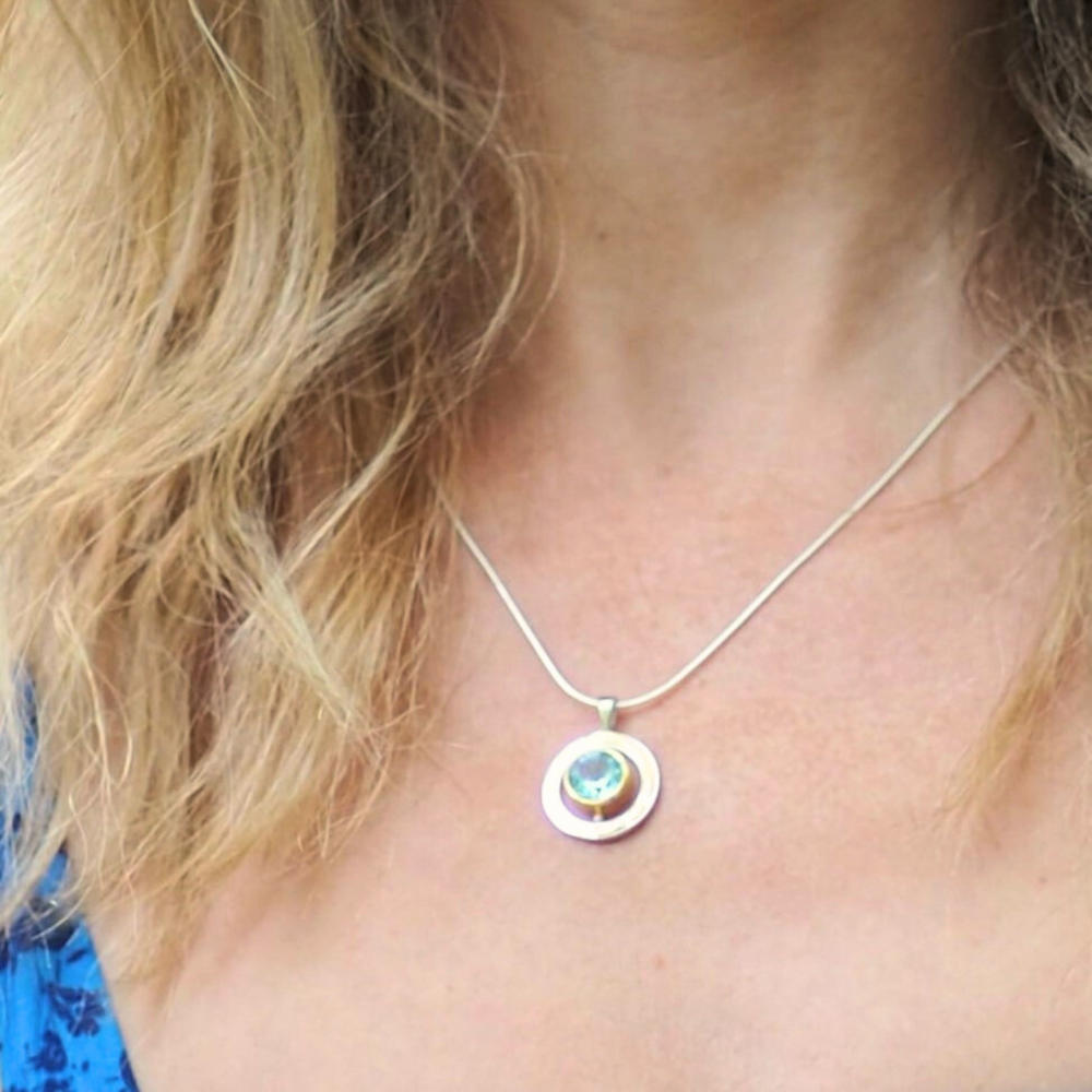 A person with wavy blonde hair wears a blue top and the Elodie Necklace by Gallardo & Blaine Designs, featuring a blue-green gemstone set in a circular silver pendant on a thin chain.
