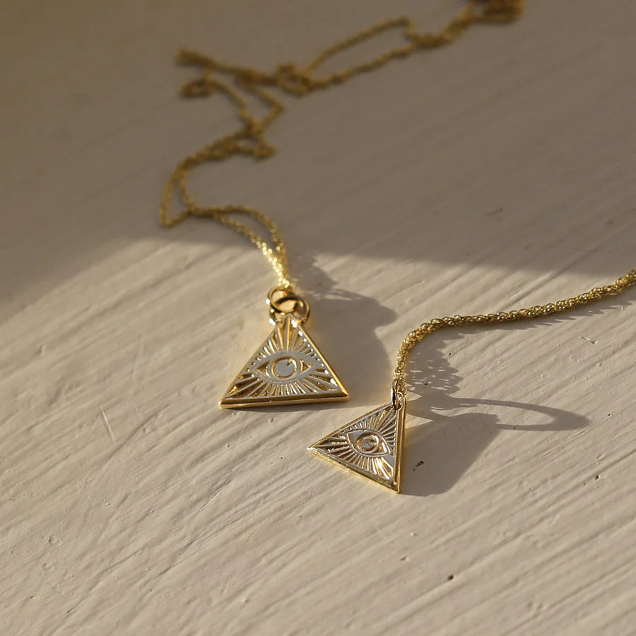 Two Little Eye of Protection Necklace Gold pieces by Andrea Mears Jewellery, each with a gold-plated triangle pendant engraved with an eye, are displayed on a light surface in gentle natural sunlight.