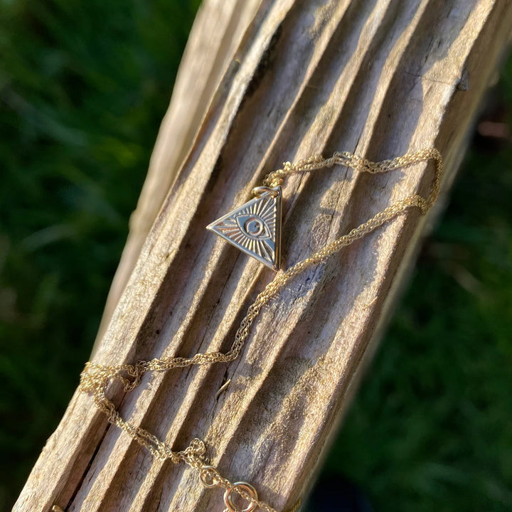 The Little Eye of Protection Necklace Gold by Andrea Mears Jewellery features a gold plated triangular pendant with an engraved eye, resting on textured wood with blurred green grass in the background.