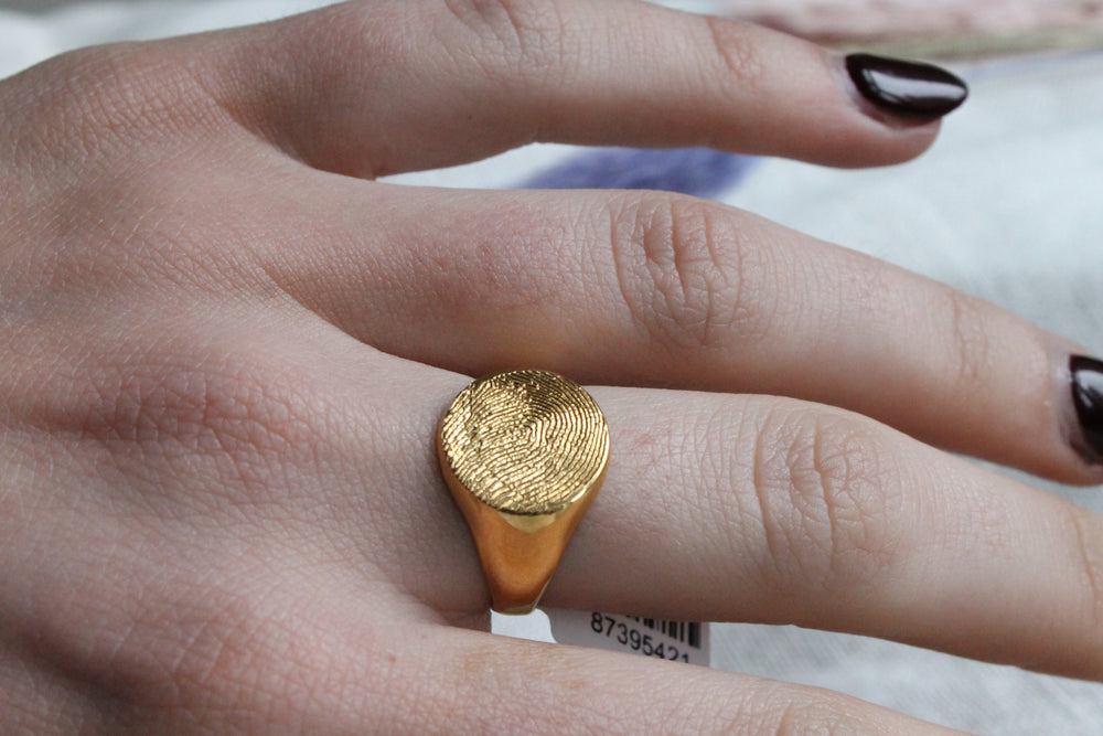 A close-up shows a hand with dark brown or black nails wearing the Personalised Charms Engraved Fingerprint Gold Plated Oval Signet Ring, featuring a raised fingerprint design.