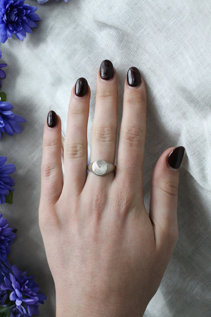 A hand with dark purple polished nails wears the Engraved Fingerprint Sterling Silver Round Signet Ring by Personalised Charms, resting on light fabric with scattered purple flowers lining the left edge.