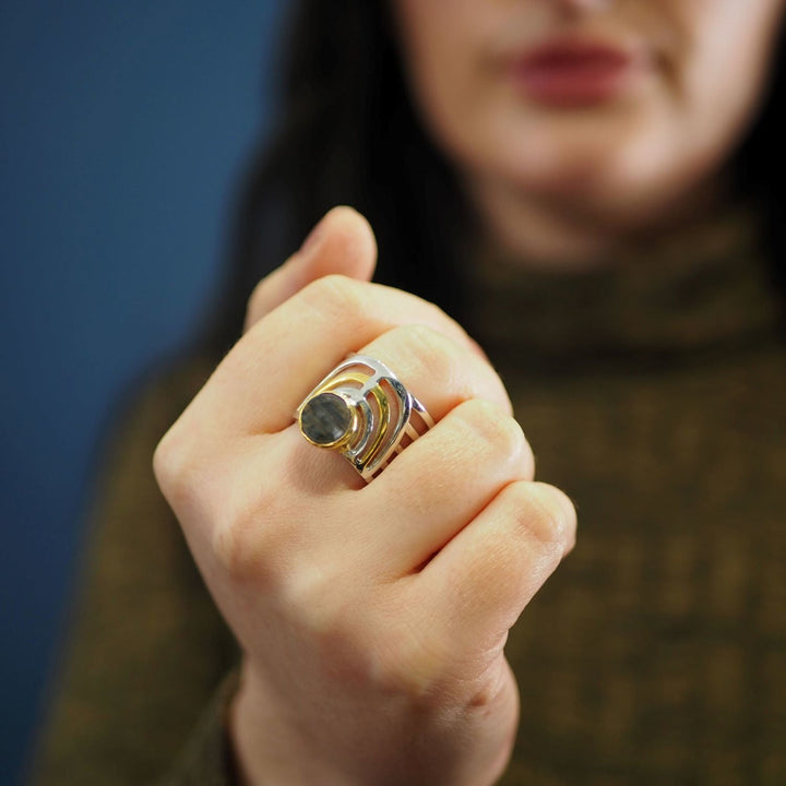 A person with long dark hair shows off the Solar Ring by Gallardo & Blaine Designs, featuring a chunky round gemstone and geometric design, against a blurred blue background.