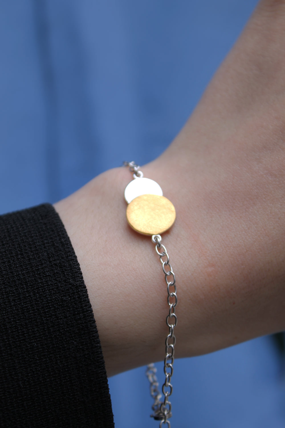A close-up shows a wrist wearing the Gallardo & Blaine Designs Archeol Bracelet in sterling silver & gold vermeil, featuring two round charms—one gold, one silver—set against blue fabric and a black sleeve.
