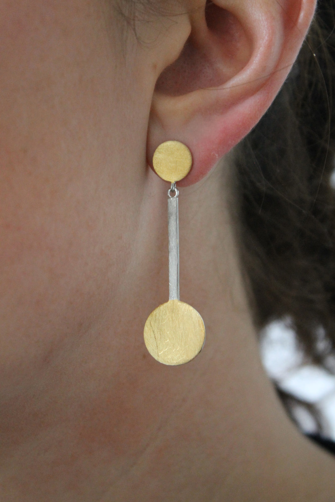 A close-up of a person’s ear wearing Gallardo & Blaine Designs’ Archeol Earrings in Sterling Silver & Gold Vermeil, with a small gold plated silver circle at the lobe, a larger gold circle below, joined by a thin silver bar.