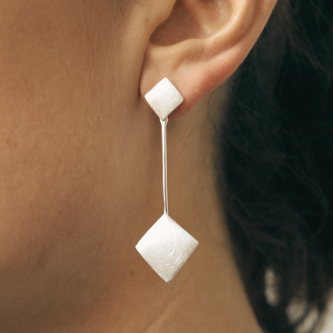 Close-up of a person's ear wearing Gallardo & Blaine Designs' Art Deco Earrings in Sterling Silver, featuring two geometric diamond-shaped pieces connected by a thin vertical bar.