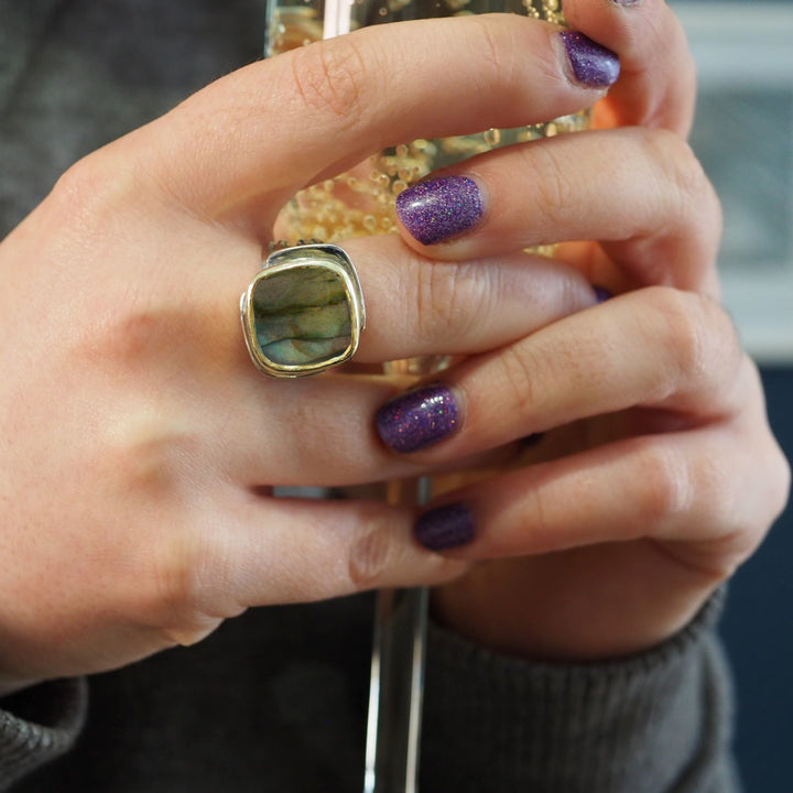 A person holds a sparkling glass with golden liquid, displaying their Adjustable Art Deco Ring in Silver Gold by Gallardo & Blaine Designs, featuring a large square gemstone. Their nails shine with purple glitter polish against an elegantly blurred background.