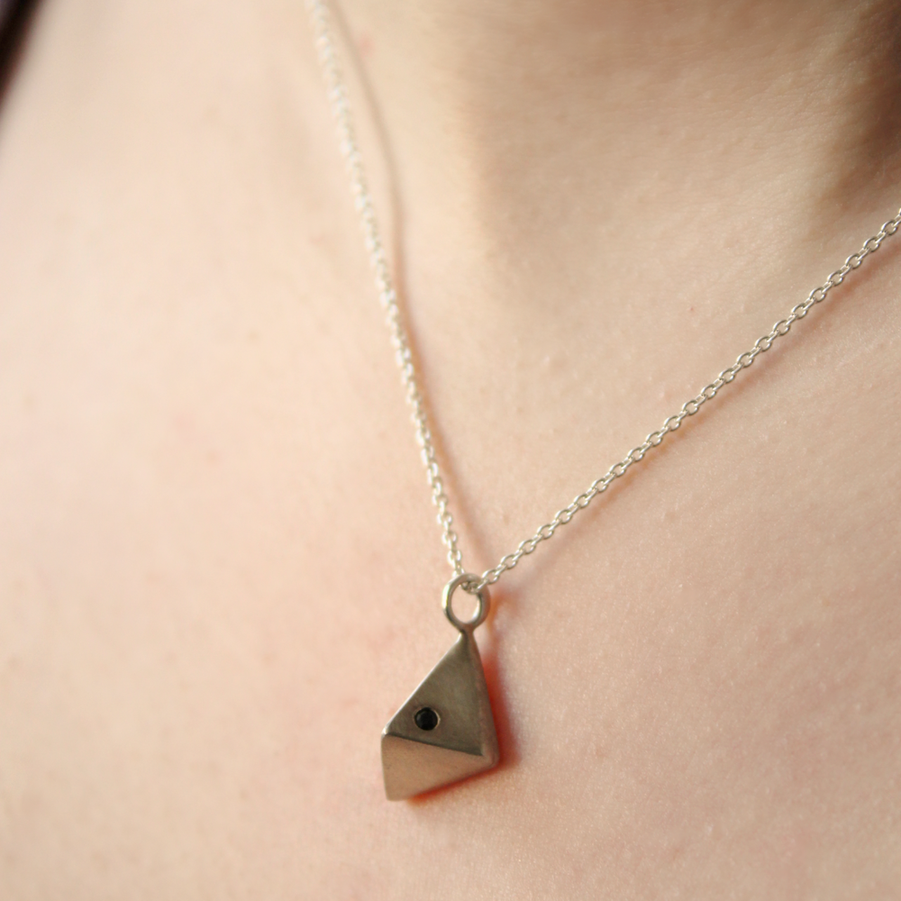 A close-up of the Atria Pendant Necklace by Gallardo & Blaine Designs—a silver chain with a small geometric triangular pendant, adorned with cubic zirconia, resting against bare skin.